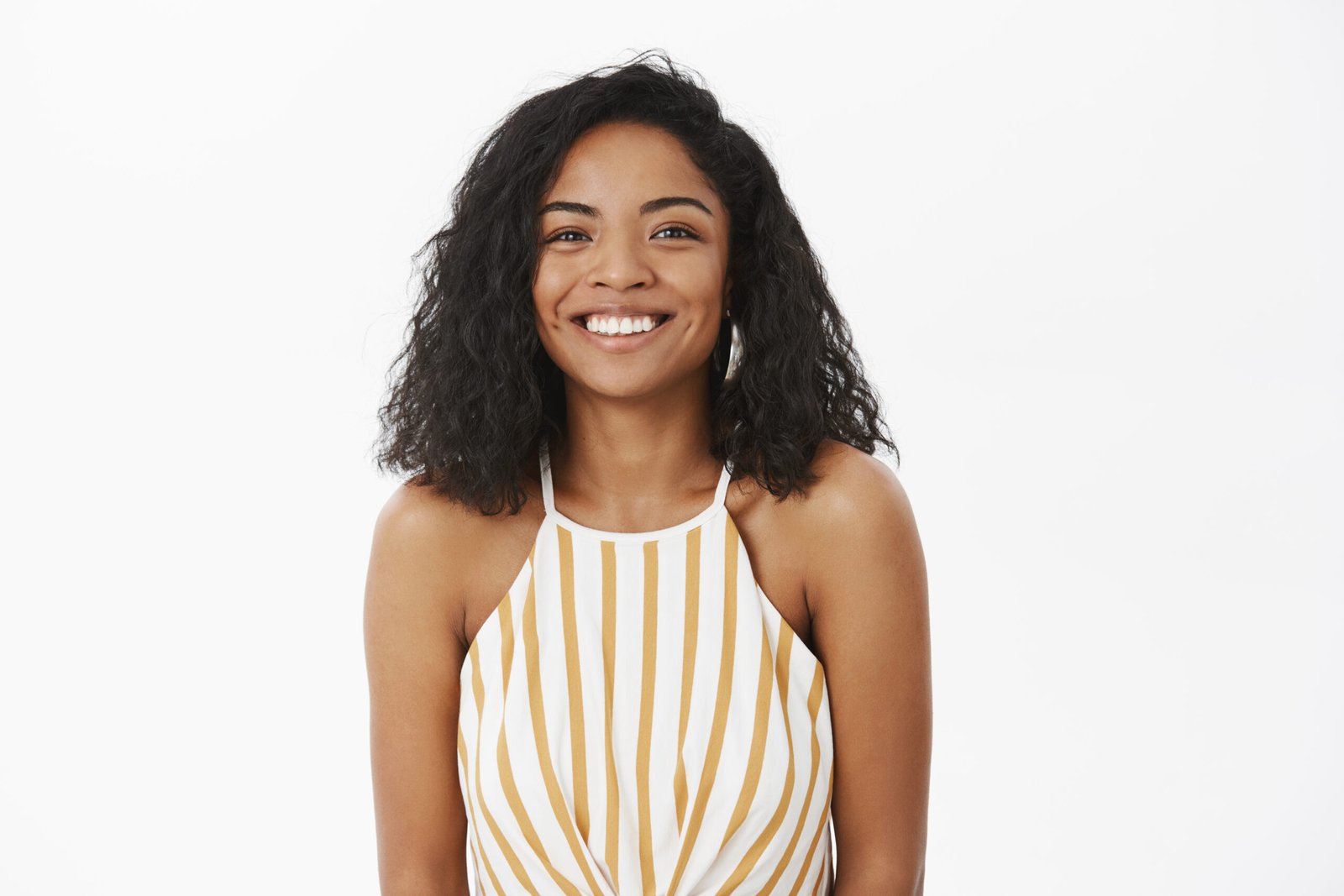 Waist-up shot of friendly-looking charming and timid african american girlfriend with curly hairstyle in trendy yellow striped top smiling from happiness and joy living perfect life over gray wall.